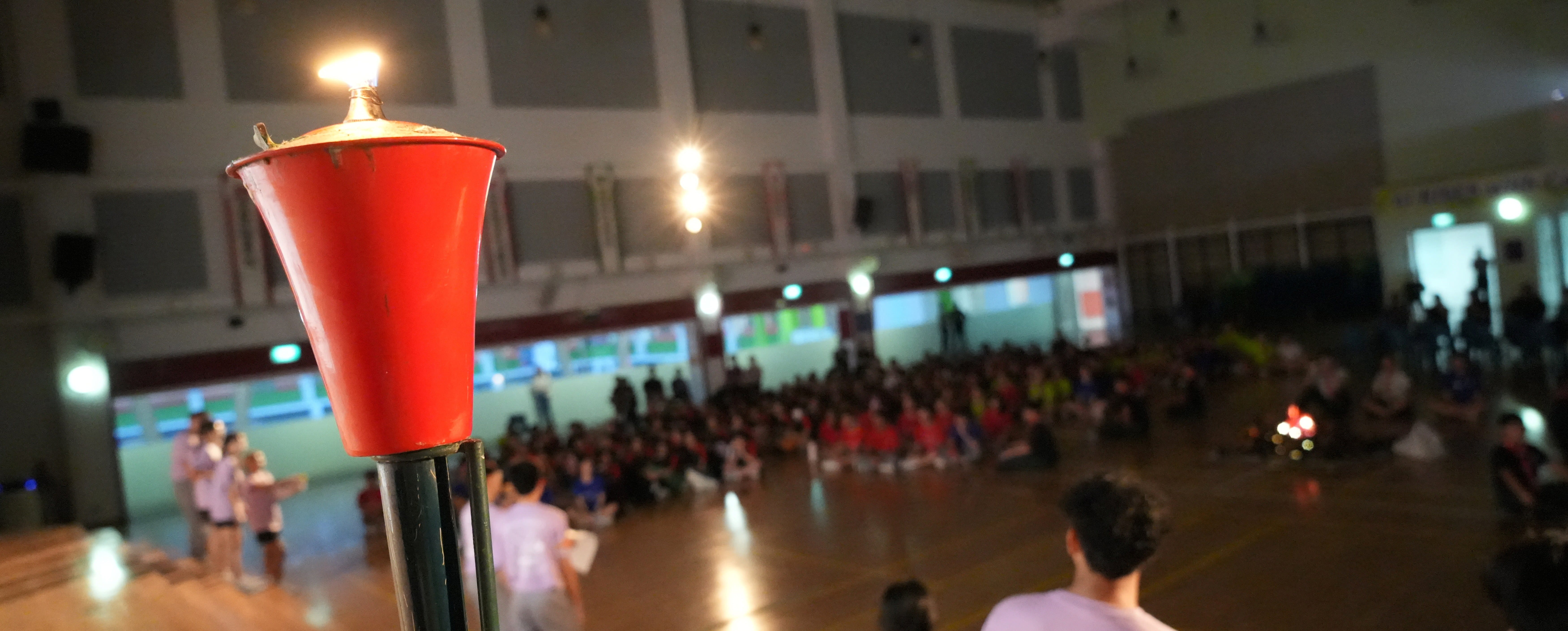 Red torch with a flame in the foreground, with a blurry crowd seated in a large indoor hall behind.